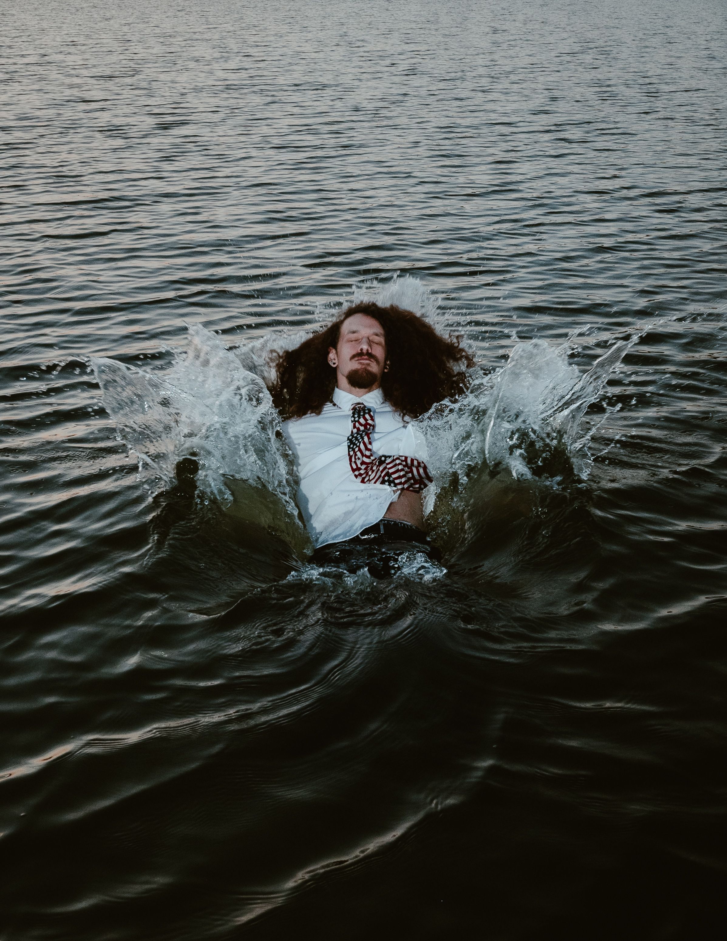 A figure creates dramatic splashes while emerging from dark rippling water on a moody overcast day.