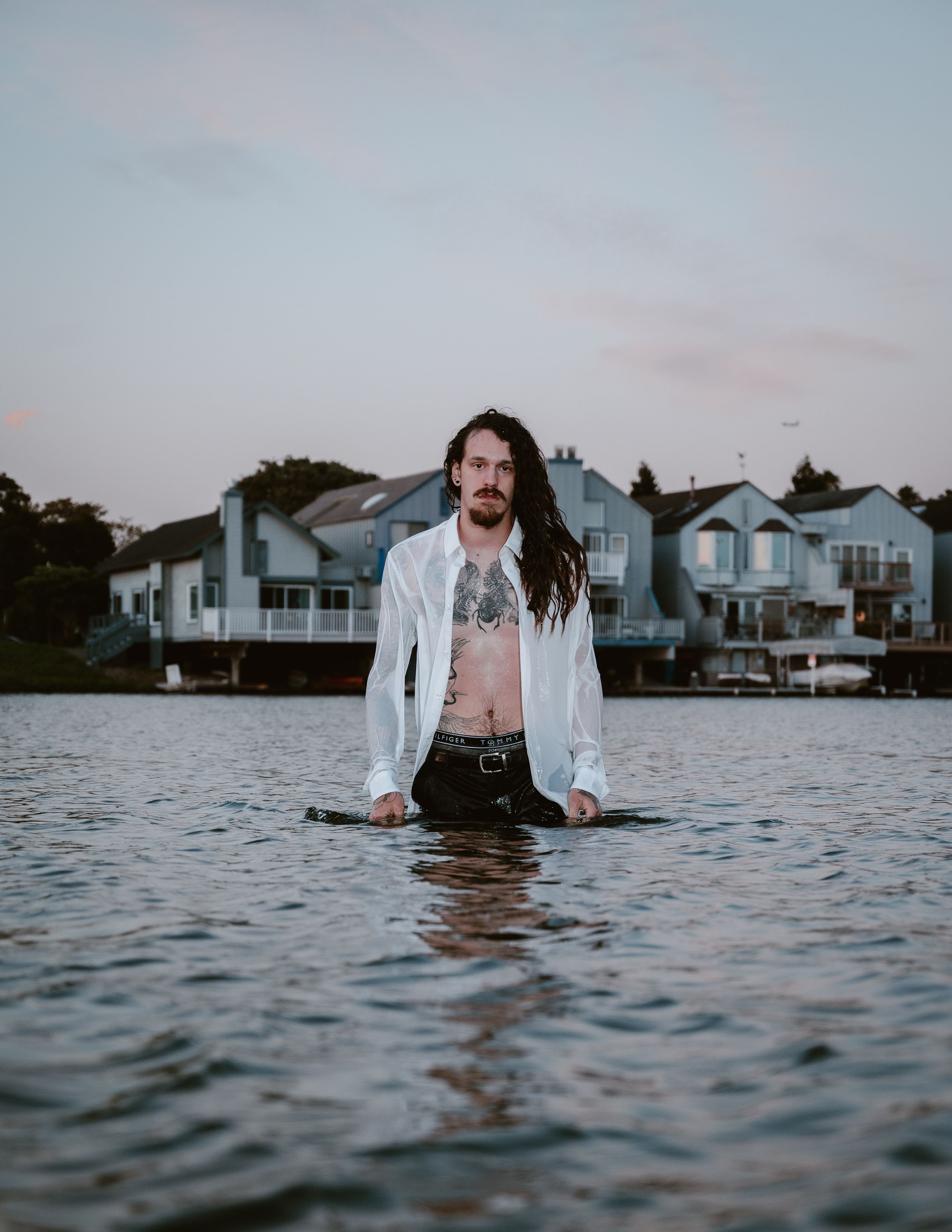 Figure standing waist-deep in water wearing white shirt with waterfront houses in background.
