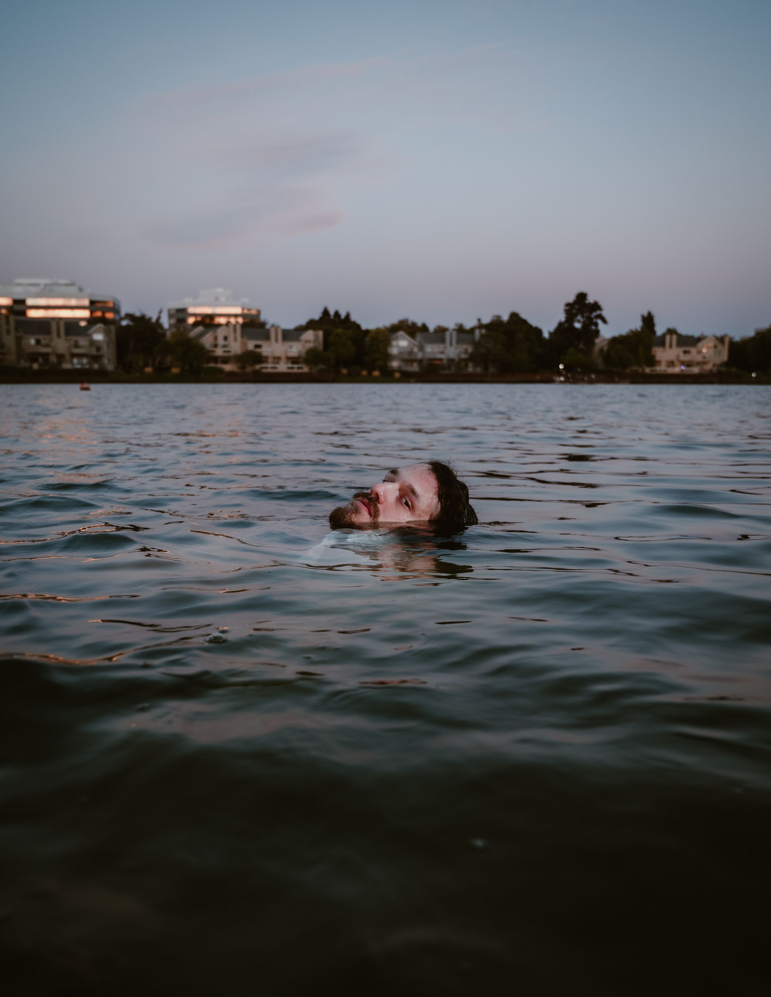 Someone floating in calm waters at dusk with lakeside houses visible along the shoreline in the background.
