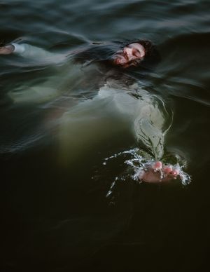 A figure floating in dark murky water creates ripples and splashes with outstretched arms in an artistic composition.