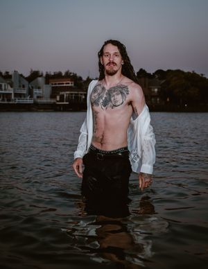 Shirtless person wearing a white open shirt stands in lake water at dusk with houses visible in background.