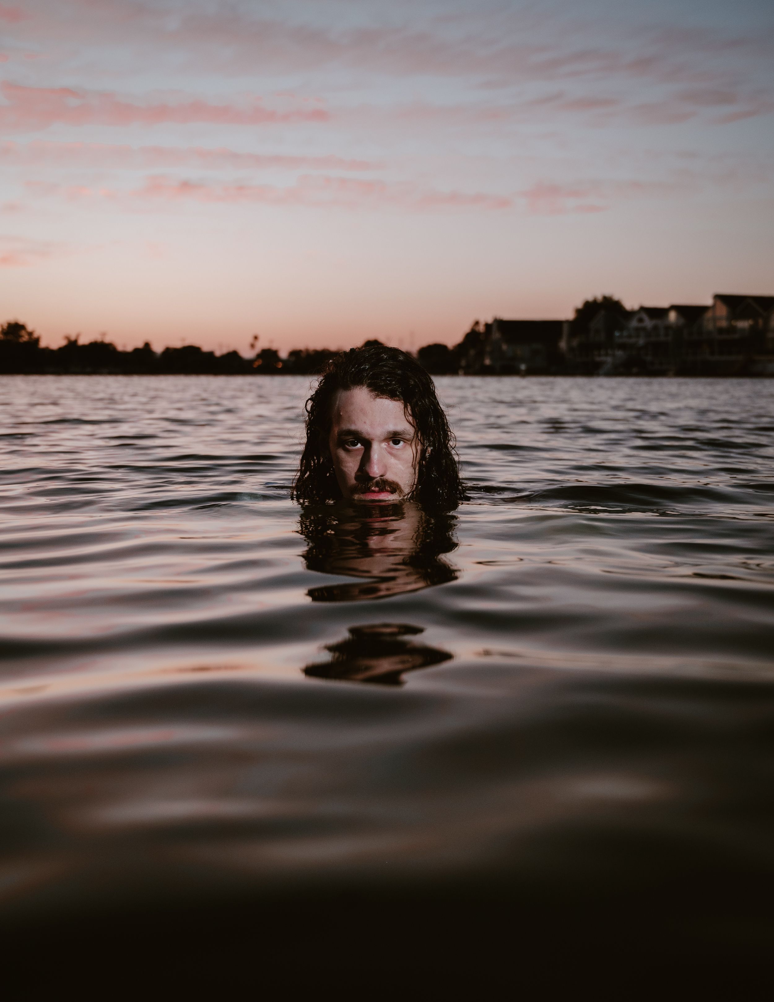 Dramatic silhouette of a person's head emerging from dark waters at sunset with buildings visible on the shoreline.