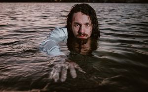 Dramatic portrait of figure partially submerged in water at dusk with rippling reflections on dark water surface.