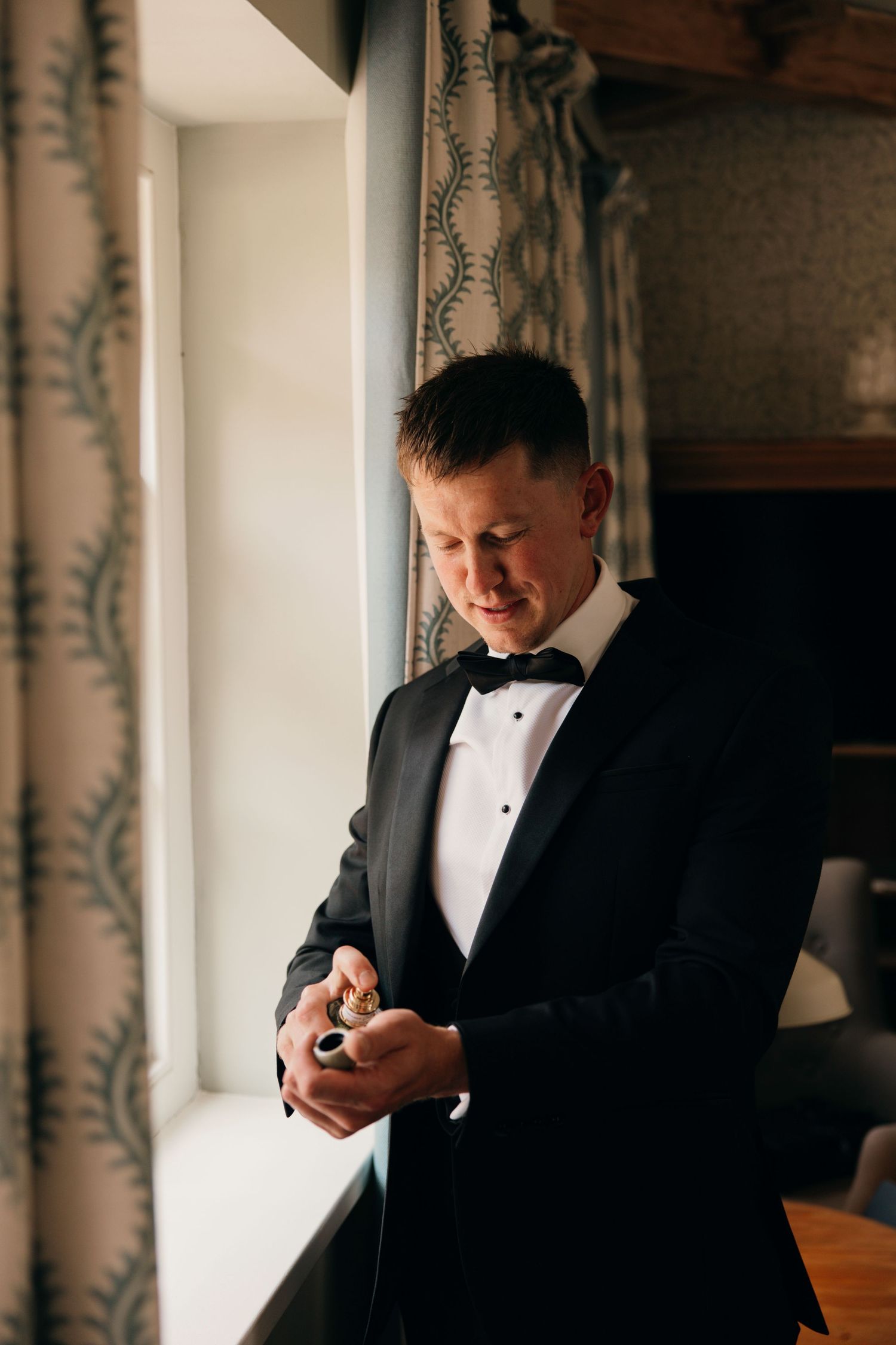 Groom in classic black tuxedo adjusts his cuffs while standing by window with patterned curtains.