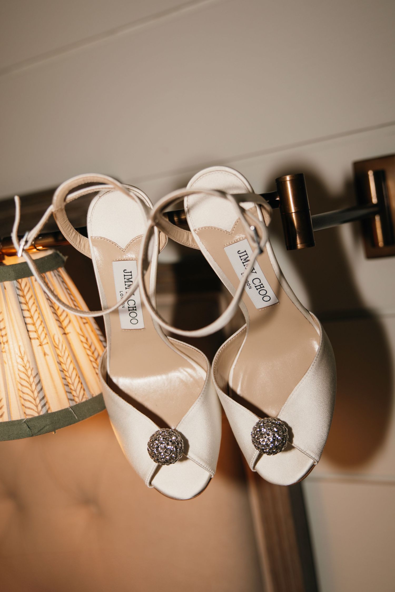 Elegant white Jimmy Choo bridal sandals with crystal embellishments hanging on a display against a warm-toned background.