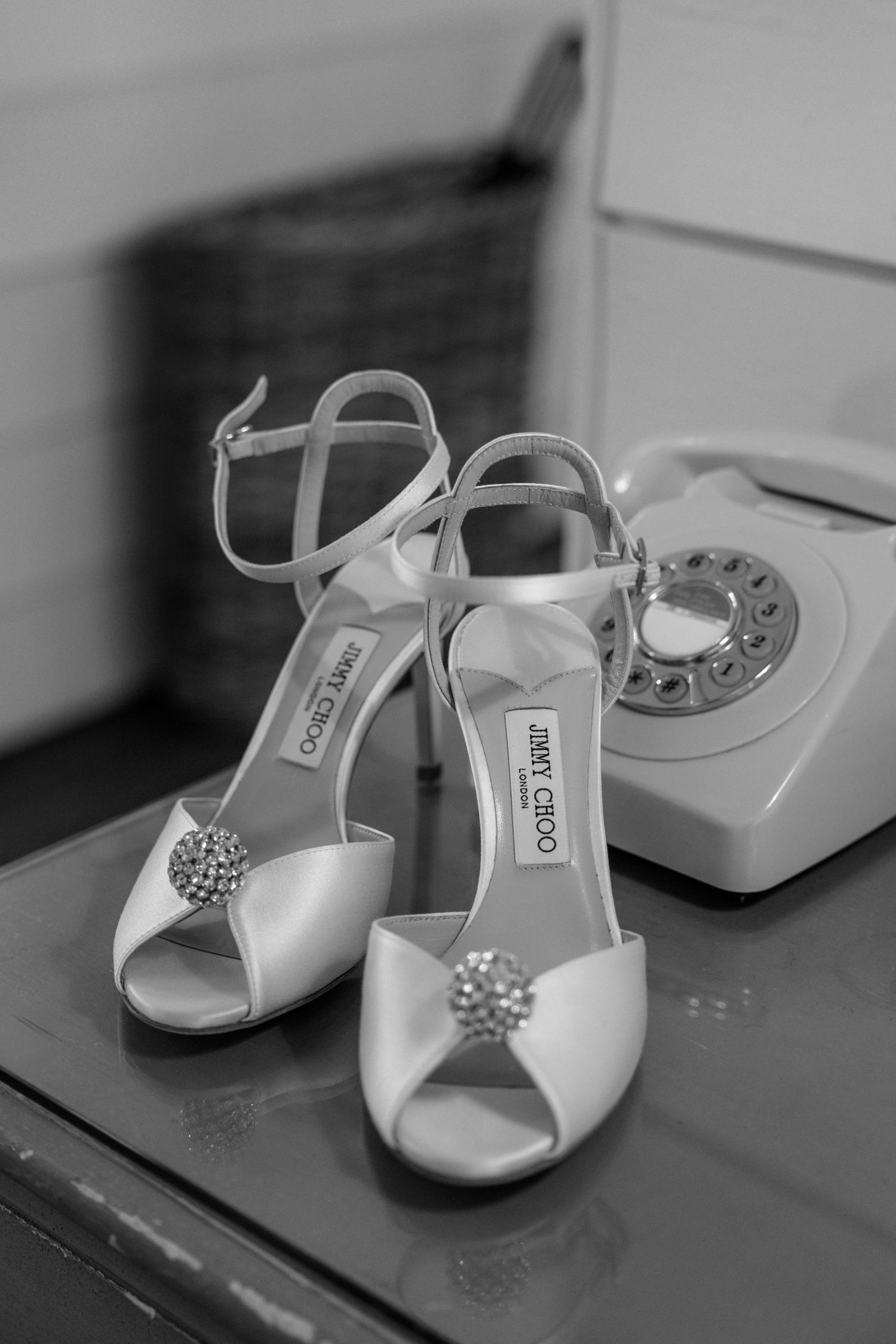 A pair of elegant Jimmy Choo bridal shoes with crystal embellishments sit next to a vintage rotary telephone in black and white.