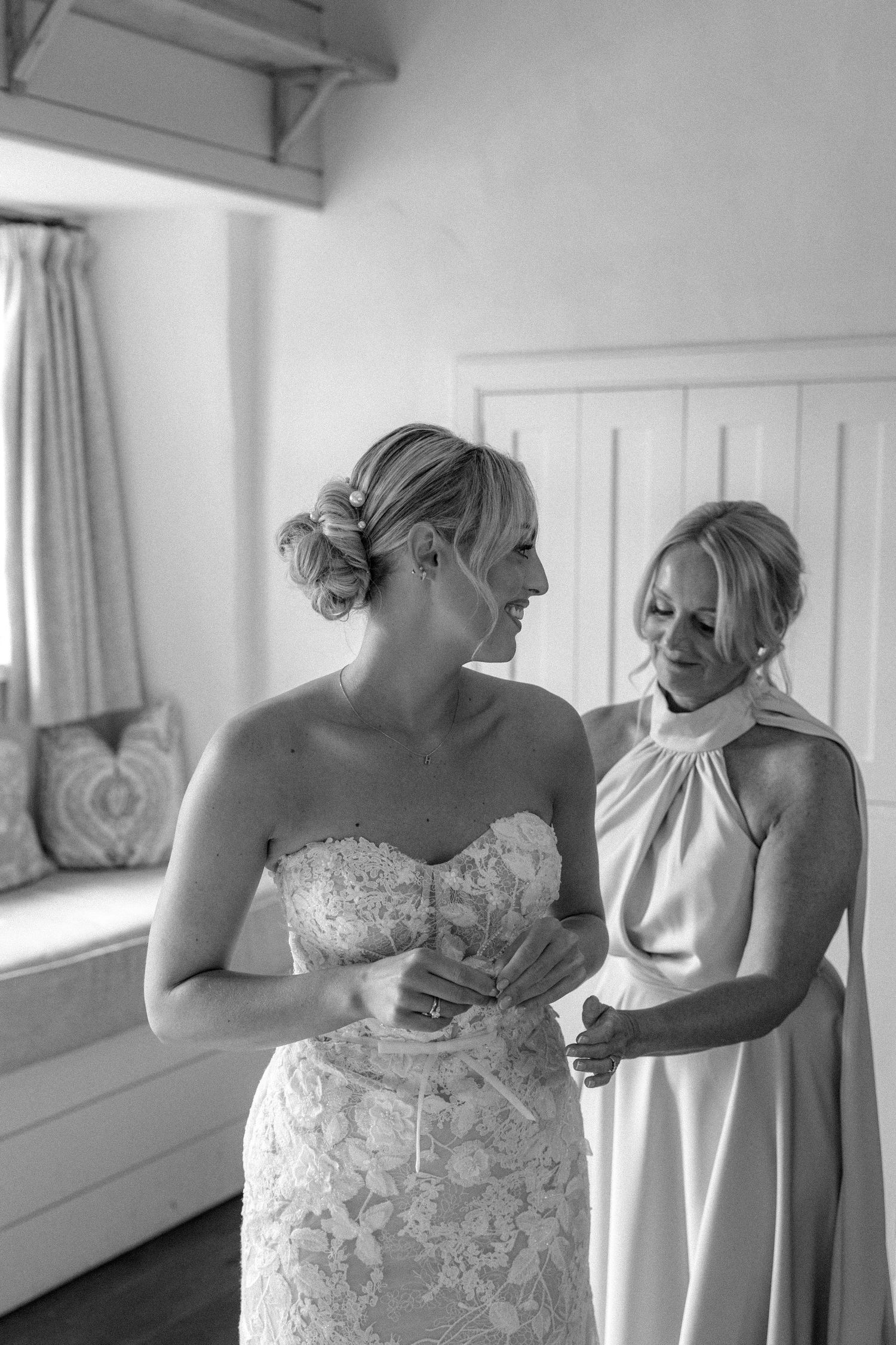 Black and white photo shows tender moment of wedding dress fitting with one woman in a strapless lace gown and helper nearby.