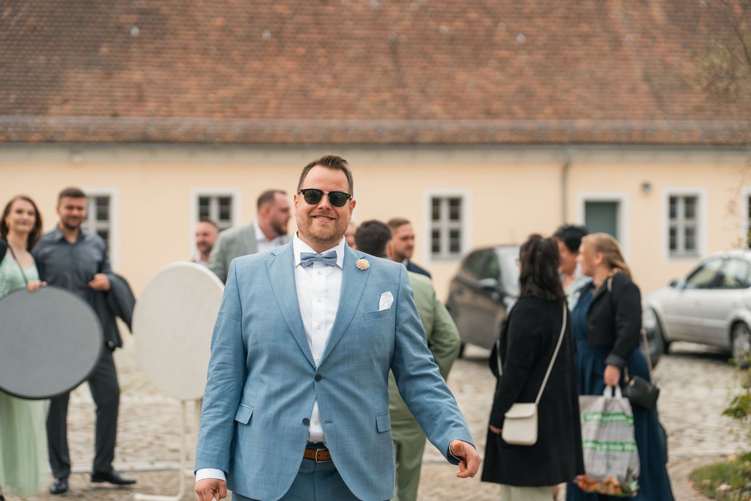 Bräutigam im blauen Anzug läuft über Hof, Gäste im Hintergrund