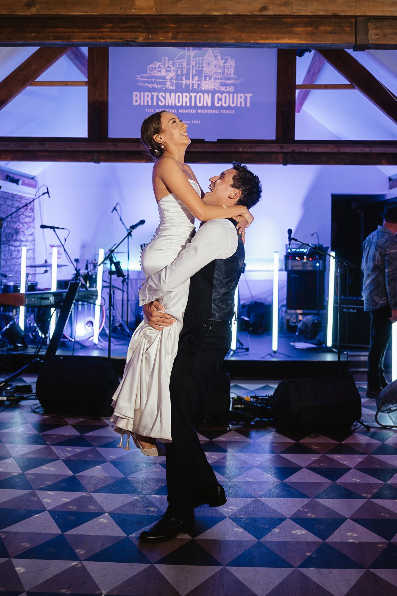 Couple shares their first dance on a black and white checkered floor under dramatic purple-lit Gothic cathedral arches.