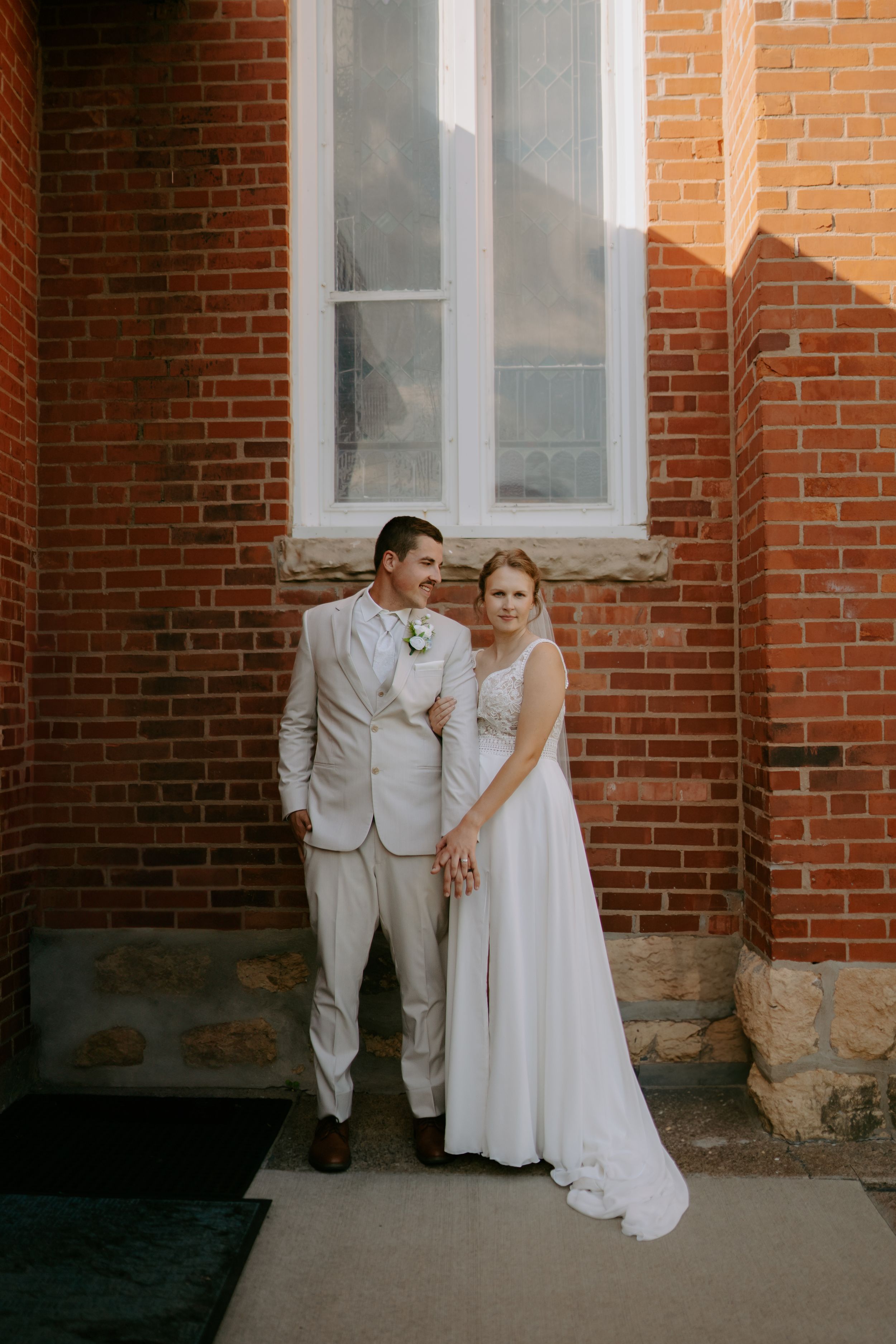 Couple poses against red brick wall in light colored wedding attire.