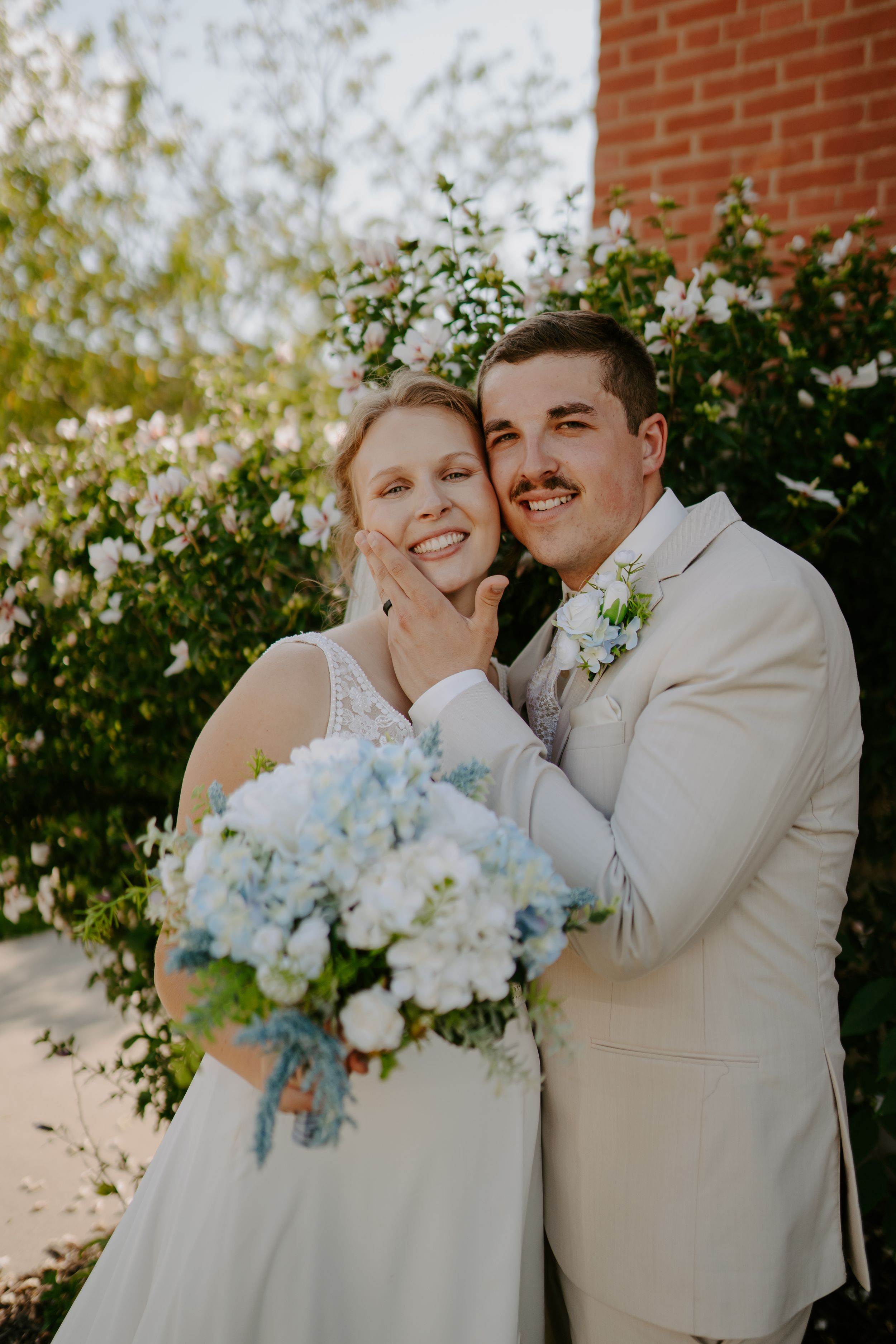 Wedding couple shares intimate moment while holding blue and white floral bouquet against brick wall.