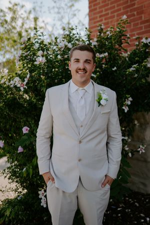A person in a cream-colored suit jacket and bow tie stands in front of a brick wall with flowering bushes.