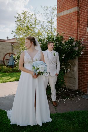 Two people in white wedding attire pose together against a backdrop of greenery and brick.