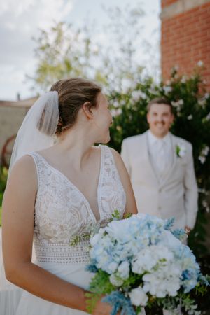 A wedding couple poses together near a brick building with white flowering bushes.
