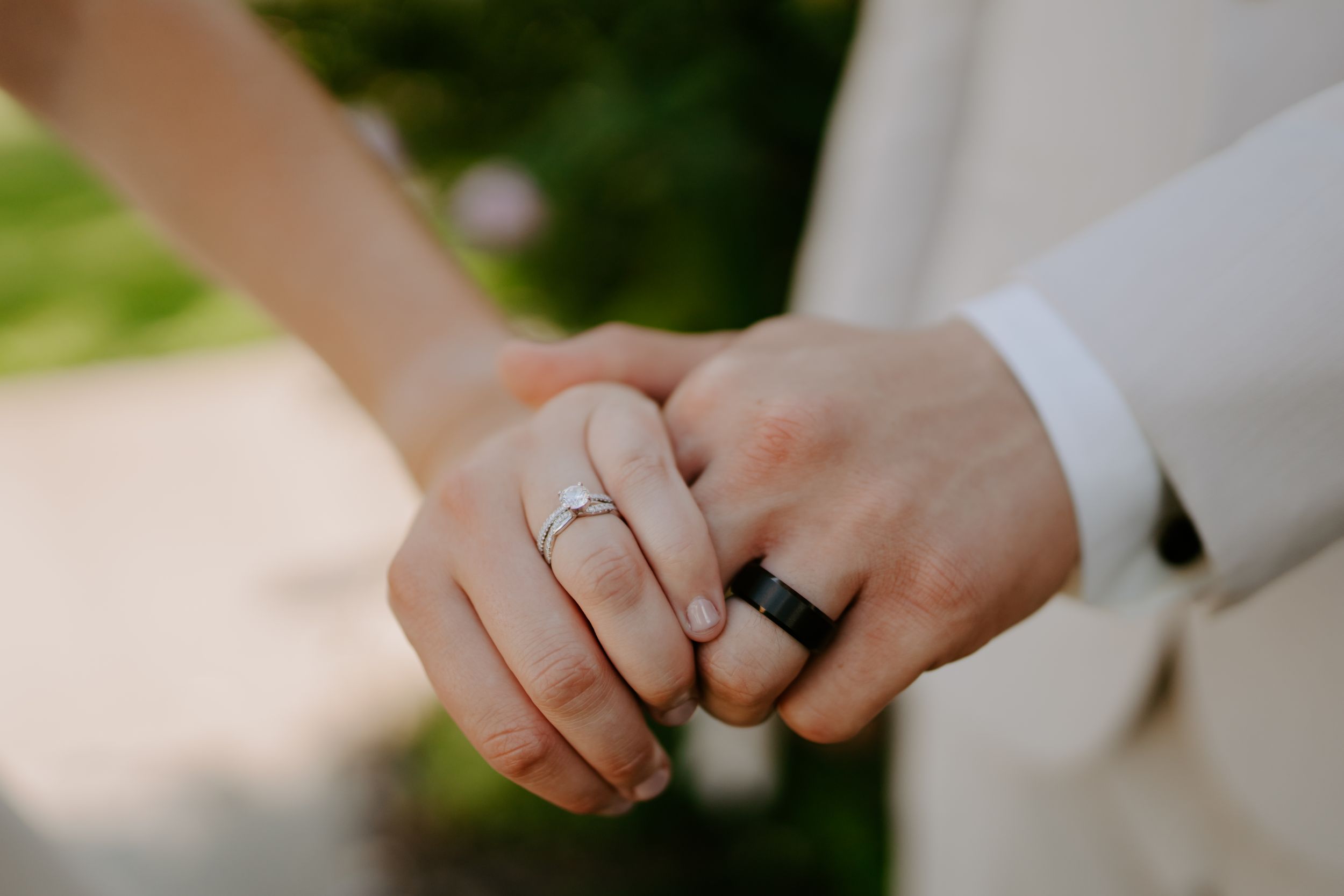 Close-up detail of wedding rings and hands against outdoor background.