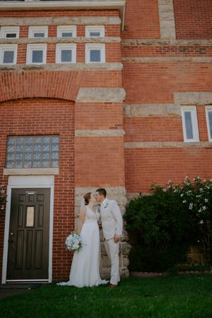 A couple in wedding attire stands together against a red brick building exterior.