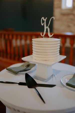 White wedding cake with a sparkly letter K topper sits on an elegant stand with serving utensils.