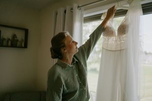 A person in grey reaches up to touch a white wedding dress hanging by a window.