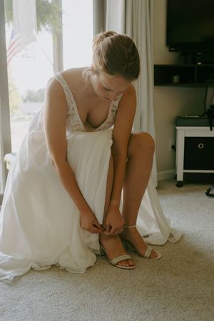 A person in white dress fastens elegant sandals while sitting in sunlit room.