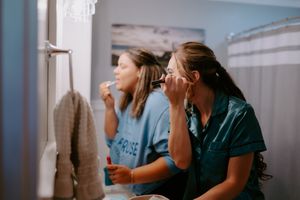 Two people doing makeup together in bathroom mirror during wedding preparations.
