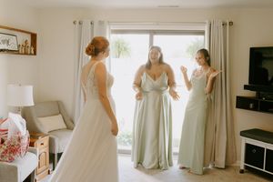 Three people in elegant dresses gather near window during wedding preparations.