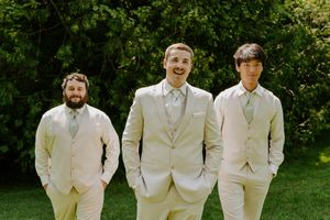 Wedding party members in cream suits pose together on grass with trees in background.
