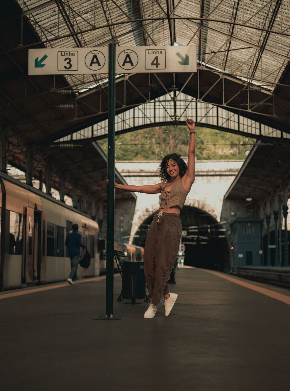 A person poses playfully under station platform signage wearing casual brown attire.