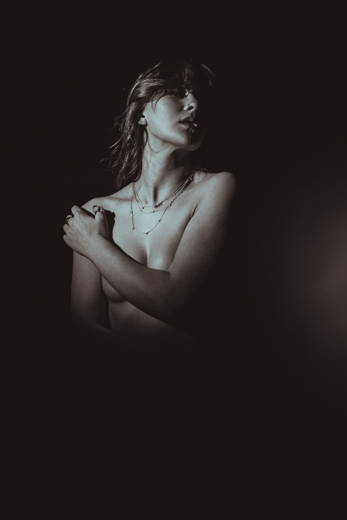 Dramatic black and white portrait of figure in shadows with necklace against dark background.