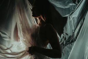 Silhouetted figure behind sheer white flowing fabric creates dramatic light and shadow effect in moody artistic scene.