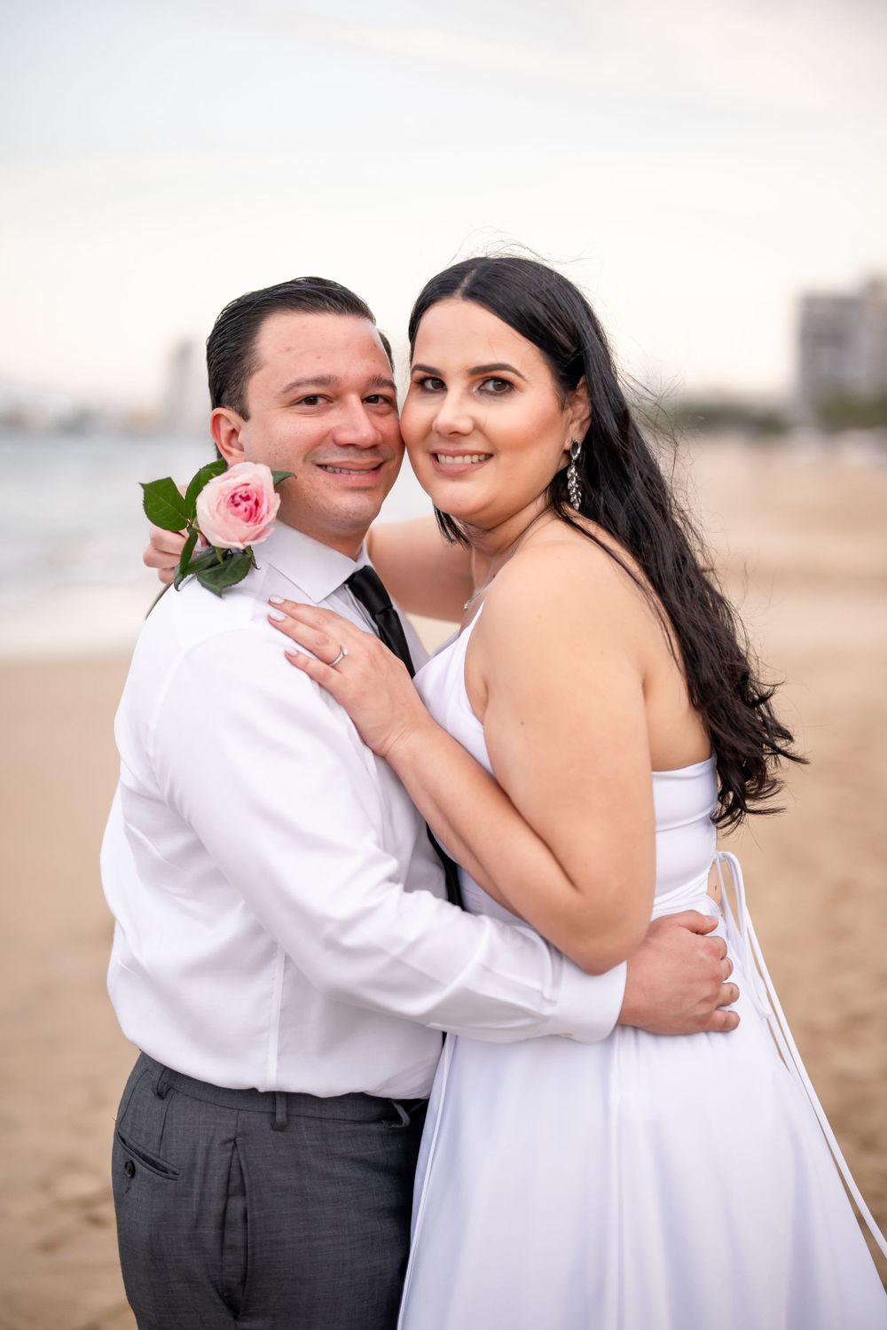 Romantic beach portrait of newlyweds with pink rose and coastal backdrop.