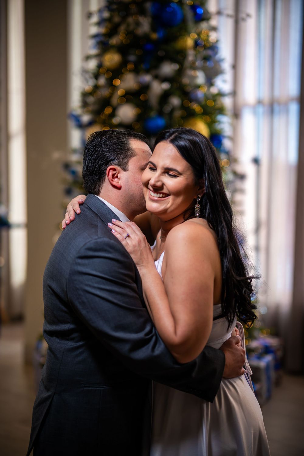 Joyful couple embracing and laughing in front of a Christmas tree decorated with blue ornaments.