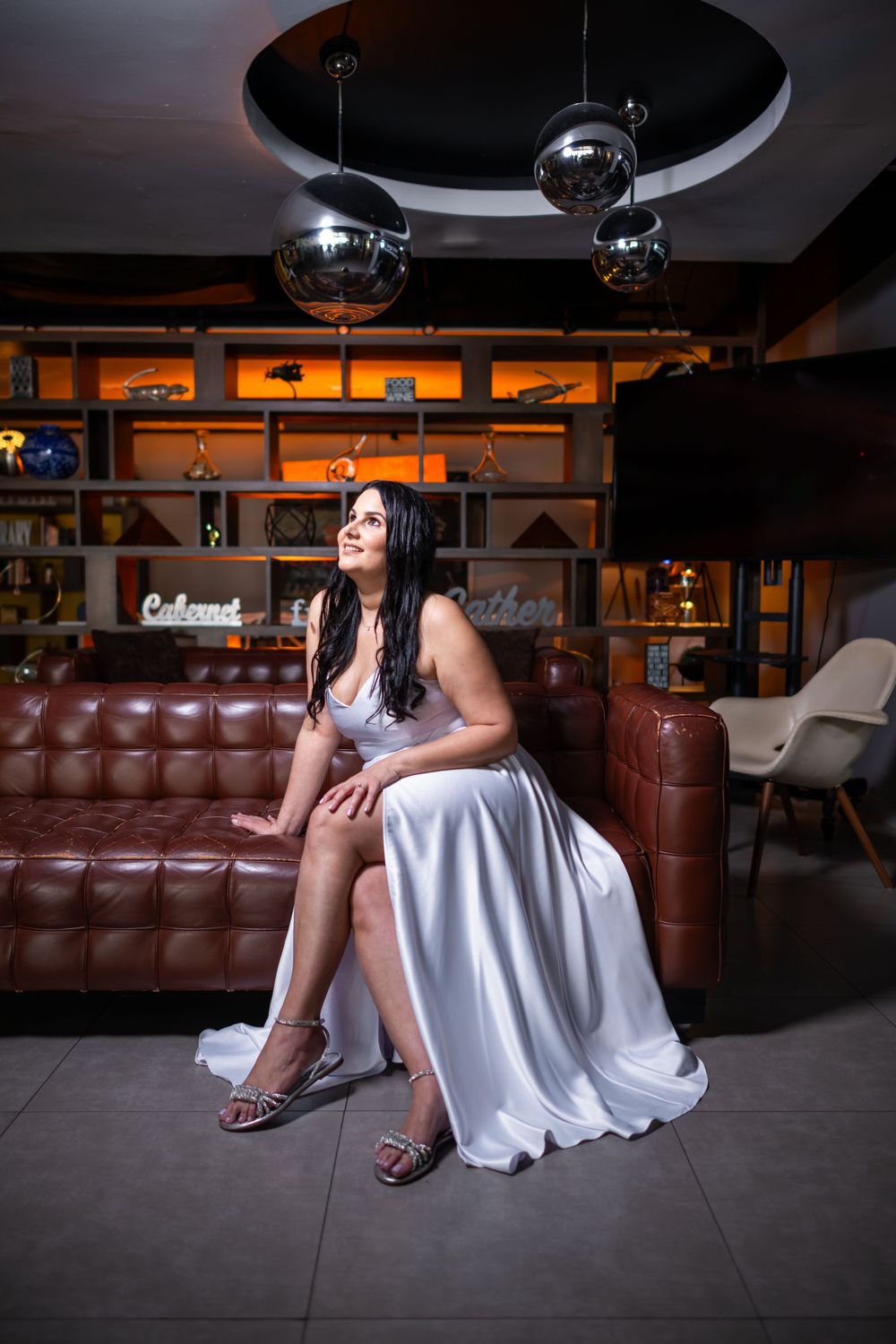 A woman in a flowing white dress sits elegantly on a leather couch in a modern lounge setting.