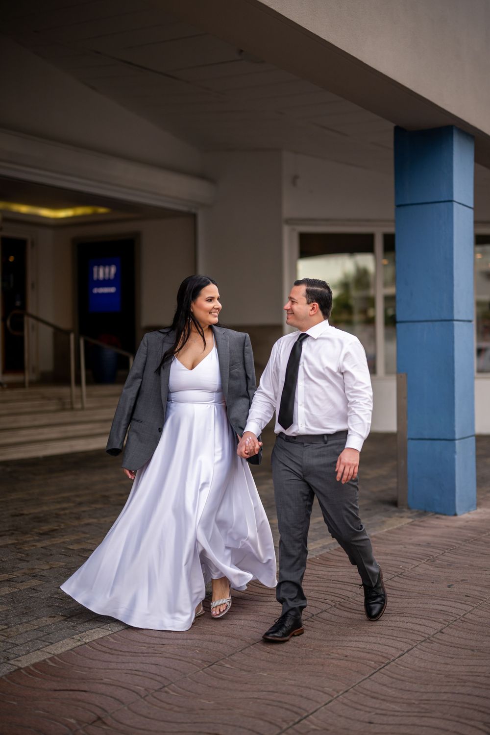 Couple strolling together outside venue, bride in flowing white dress and groom in business attire.