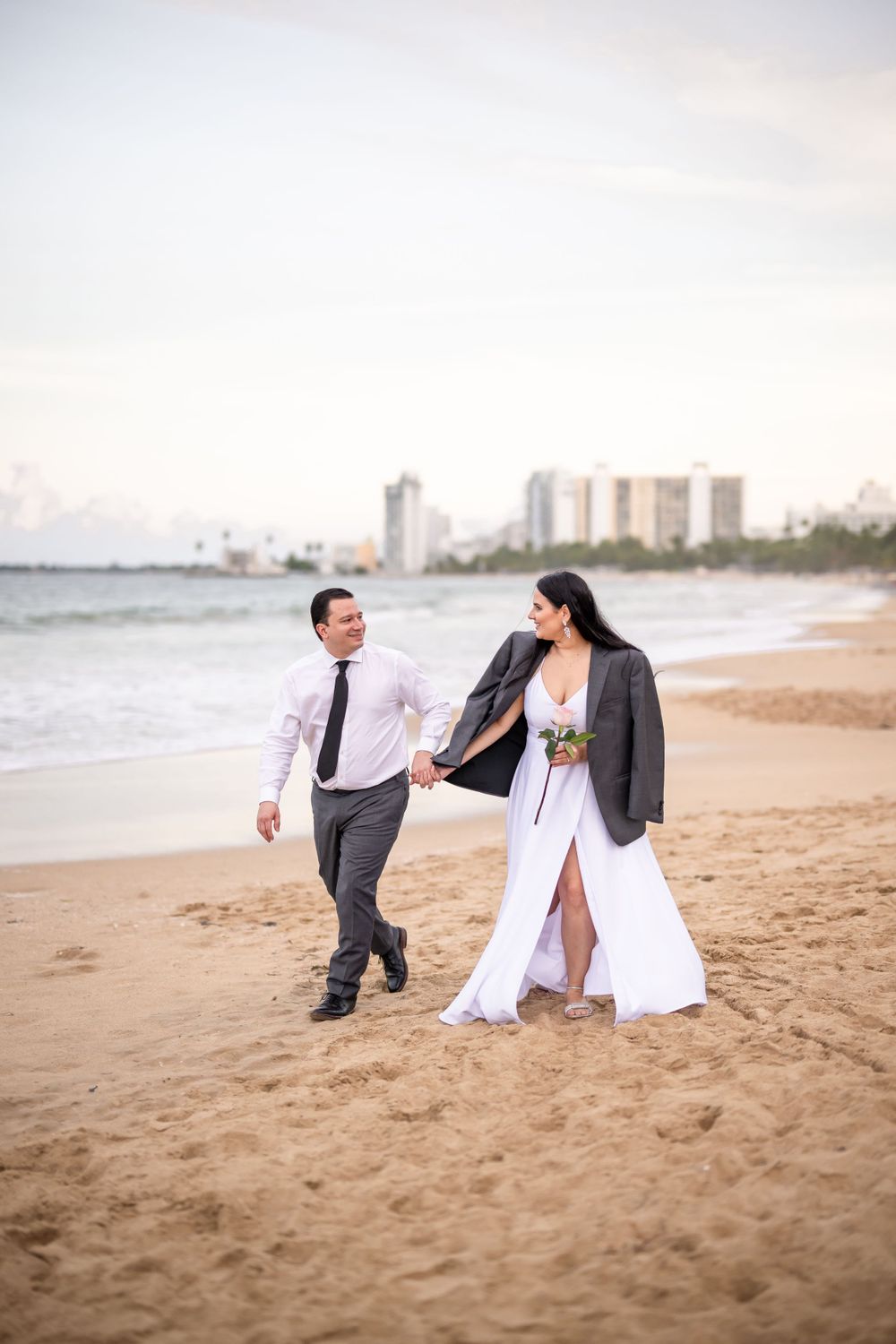 Couple walking hand in hand on beach with cityscape in background at dusk.