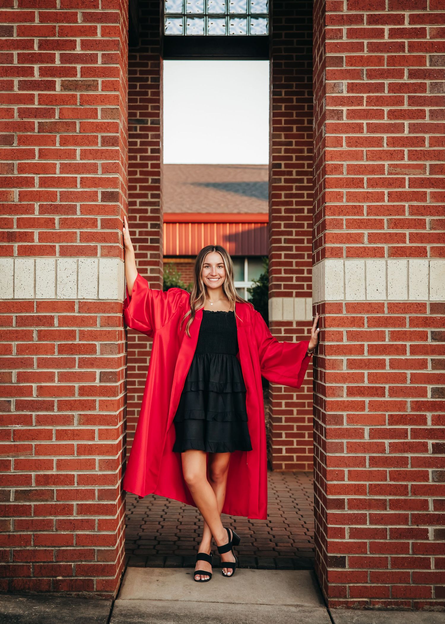 A person poses against red brick wall wearing black dress and red jacket.