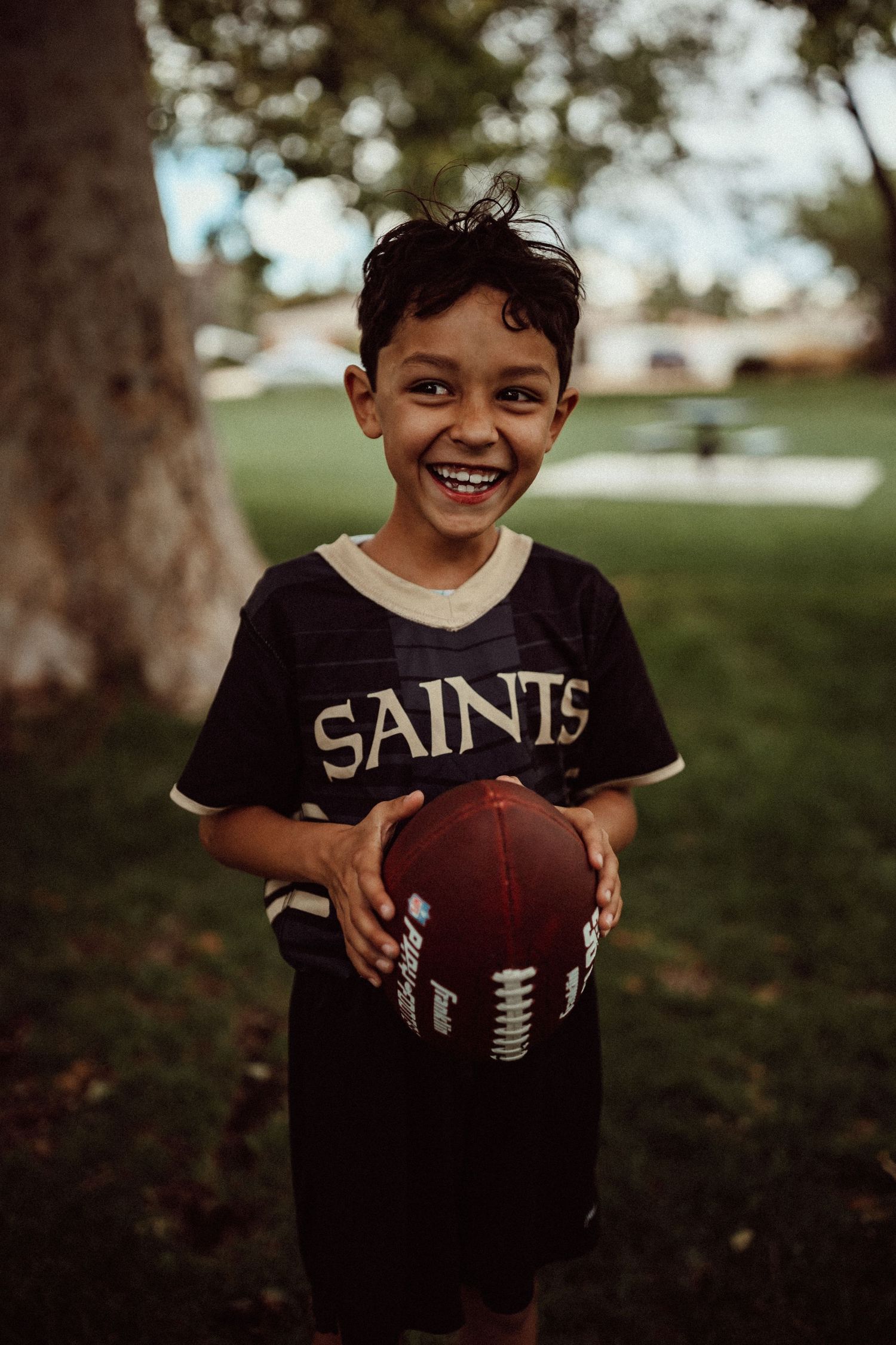 Young sports fan in Saints jersey holds football while smiling outdoors.
