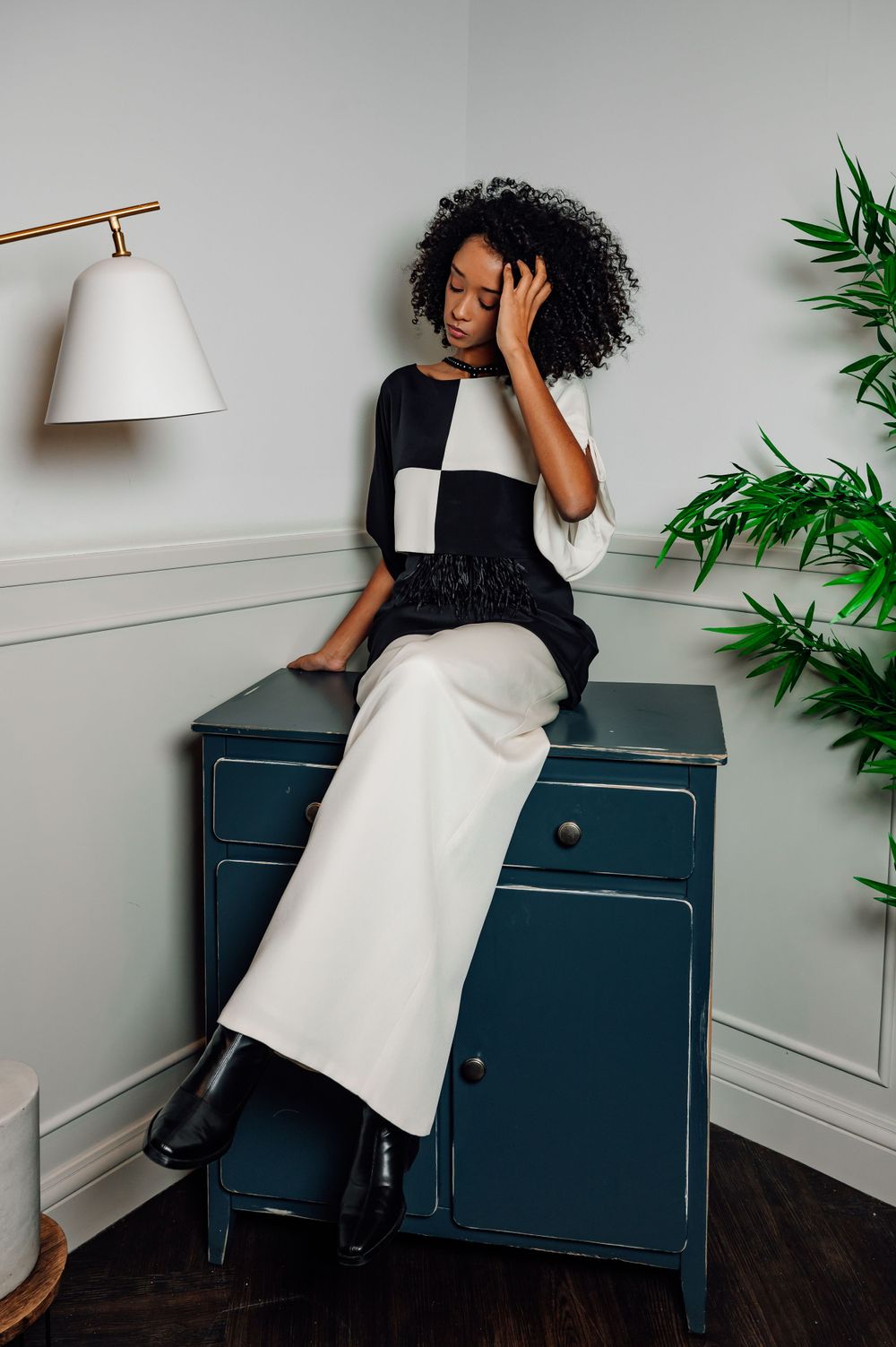 Fashion model in black and white outfit poses artistically on blue cabinet against grey wall with green plant decor.