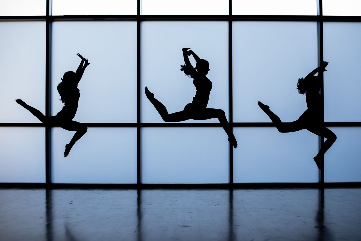 Three silhouetted figures leaping gracefully across backlit window panels.
