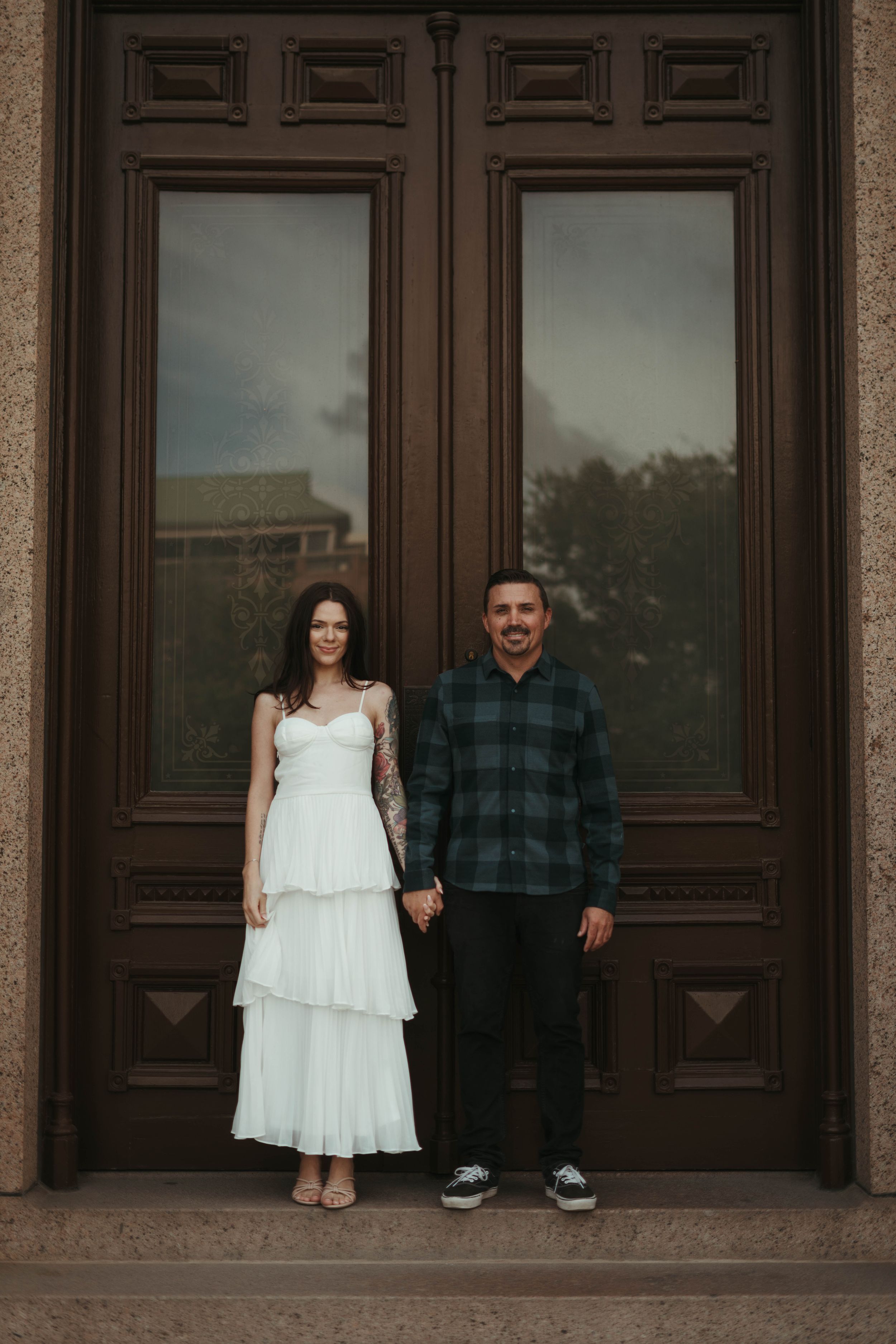 Couple poses in front of tall wooden doors at historic stone building.