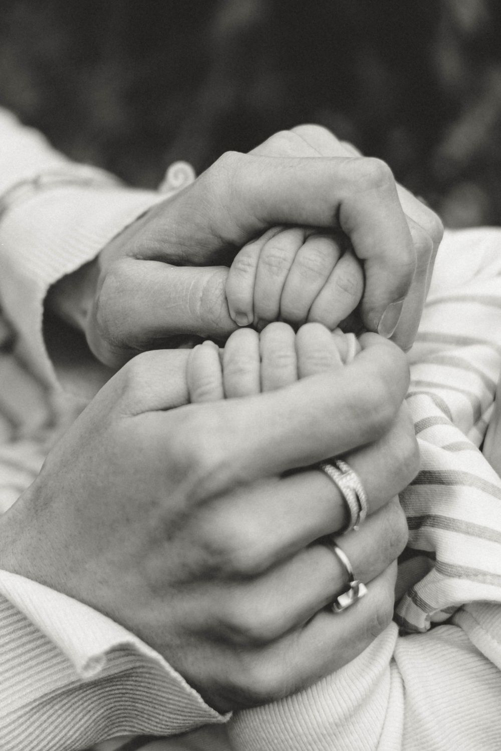 Black and white close-up of hands holding baby's tiny fingers with wedding rings visible.
