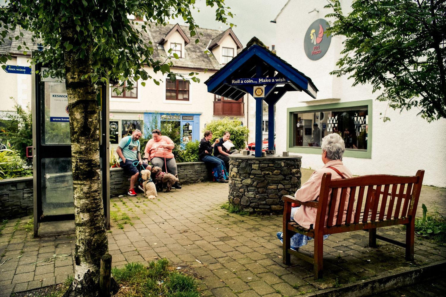 People resting at a covered bus shelter with stone walls and wooden benches in a quaint village.