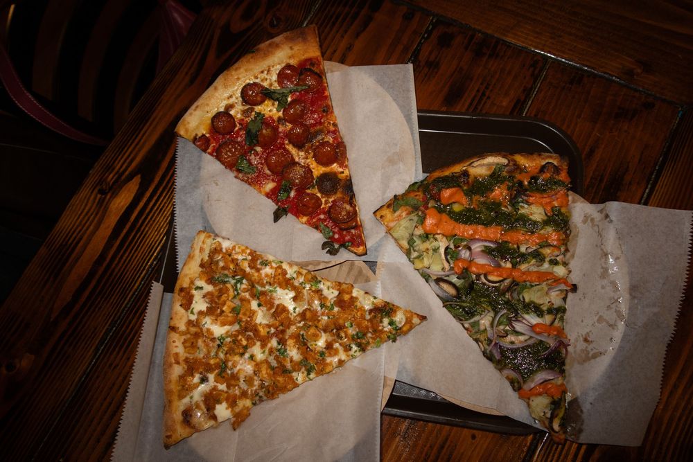 Three different slices of gourmet pizza with various toppings served on white paper on a dark wooden table.