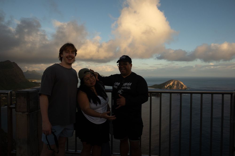 Three people pose together on an outdoor lookout point at sunset with dramatic clouds and ocean views in the background.