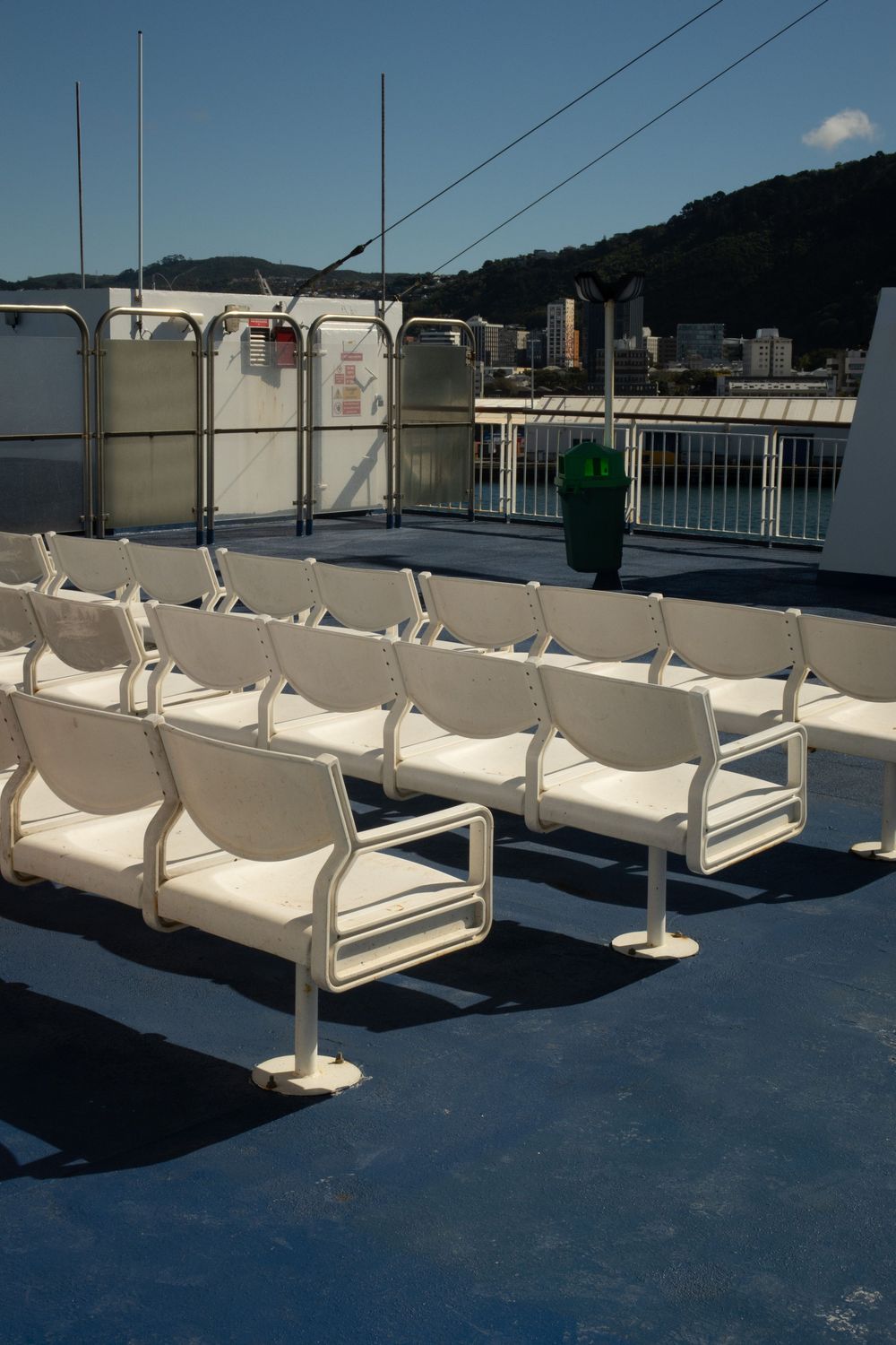 Rows of white plastic seats mounted on a blue deck floor with storage tanks visible in the background on a ferry.