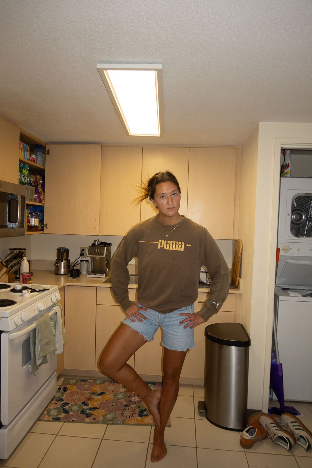 Person posing casually in a small apartment kitchen wearing brown sweatshirt and denim shorts.