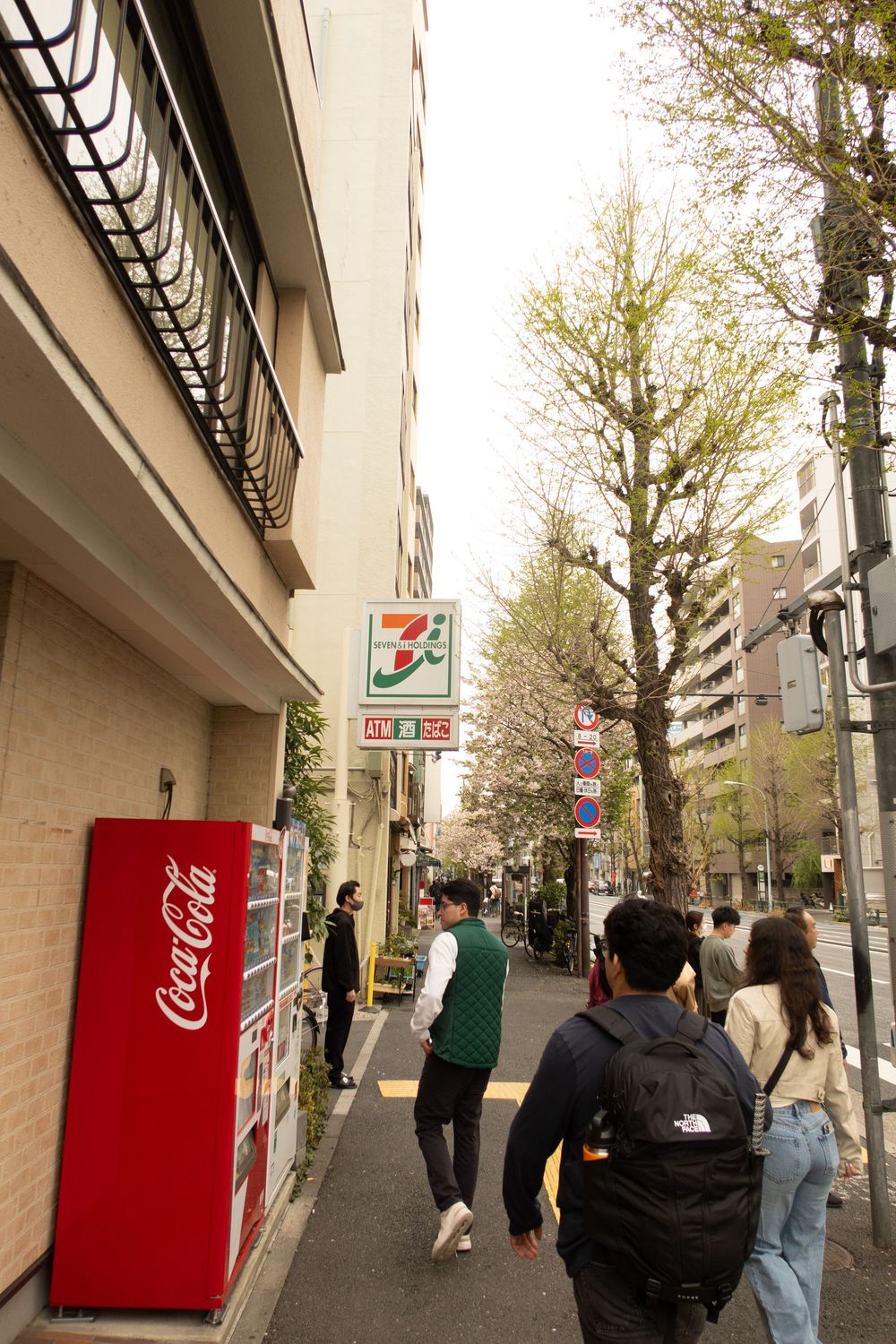 People walking on a Tokyo street with a red Coca-Cola vending machine and 7-Eleven store sign visible among spring trees.