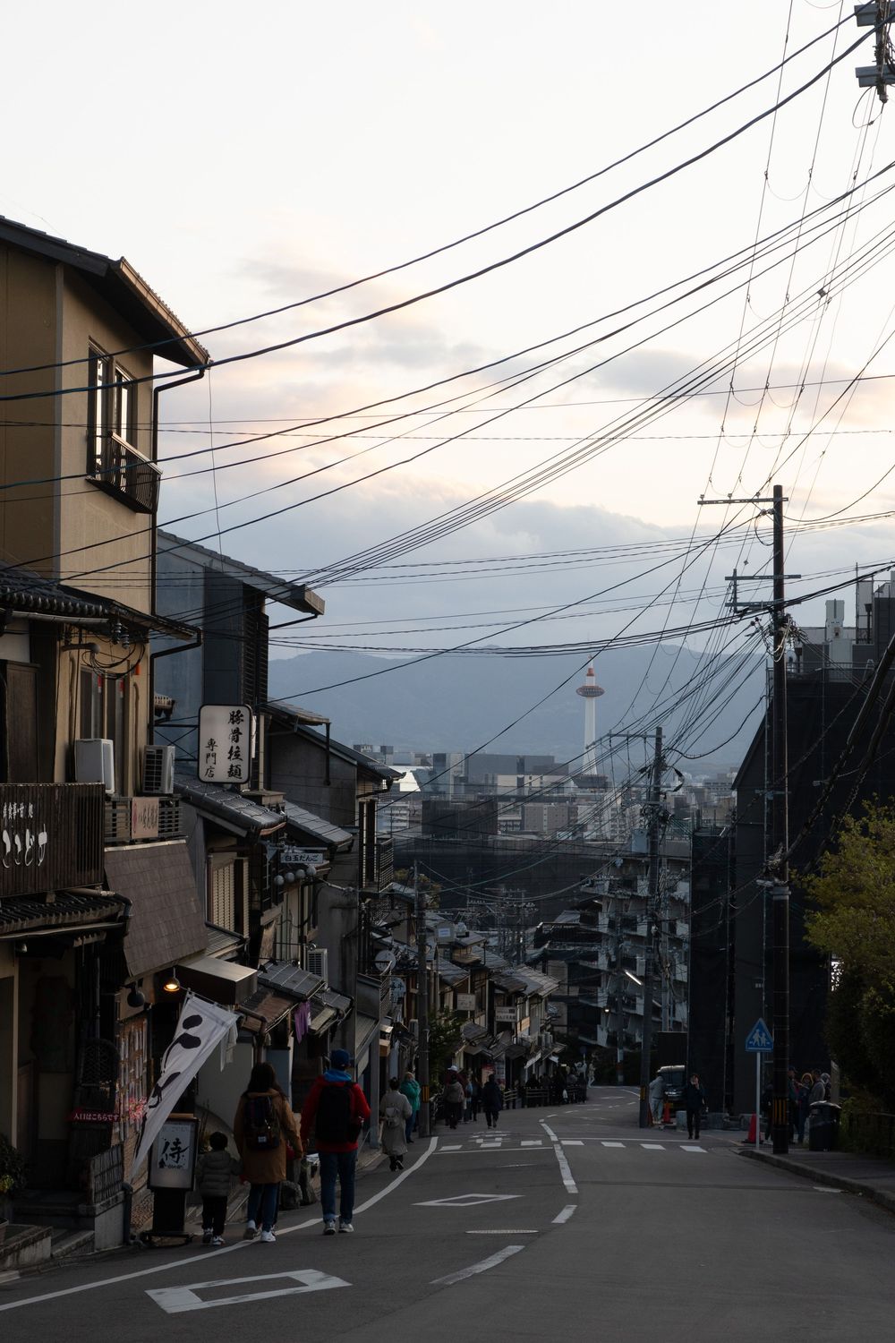 Steep residential street in Japan with power lines and traditional buildings leading downhill at dusk.