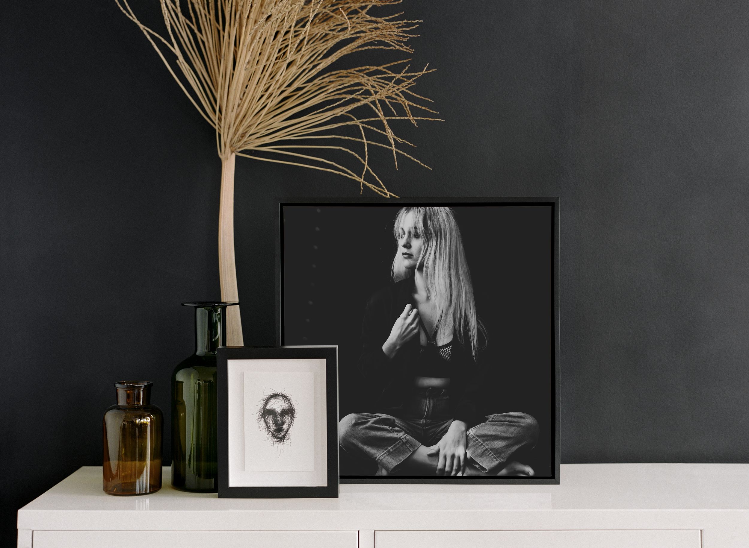 Black and white portrait photographs and dried pampas grass displayed on white surface against dark wall.