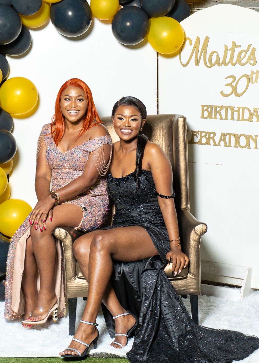 Two people sitting in gold chair at formal birthday celebration with balloon decor.