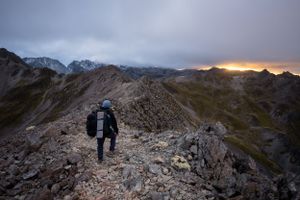 Hikers traversing a rocky mountain ridge with dramatic clouds and peaks in the background.